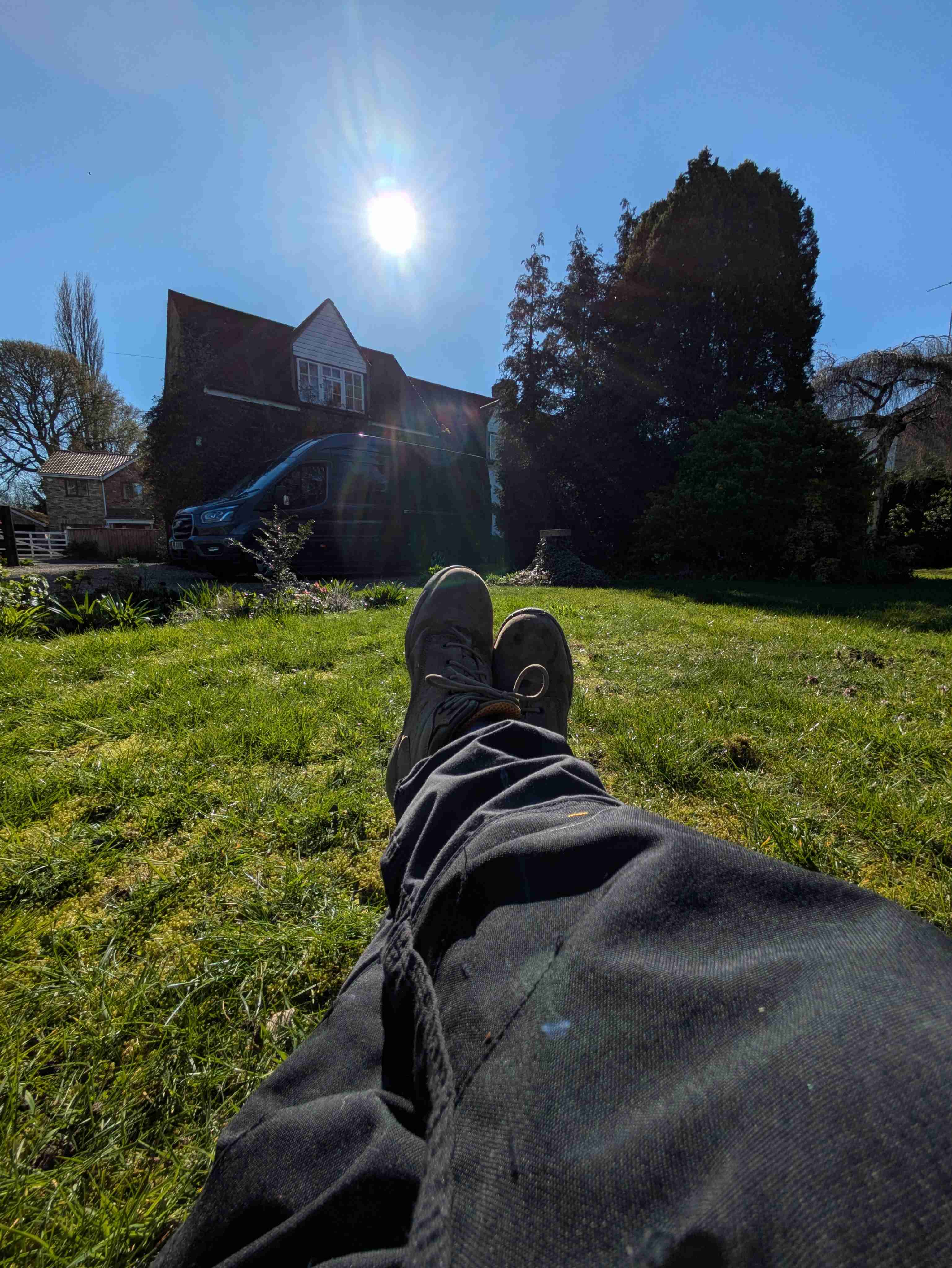 Looking down across a pair of work boots on bright spring grass, the day's job — a Cotswold-stone house — silhouetted in the sun behind