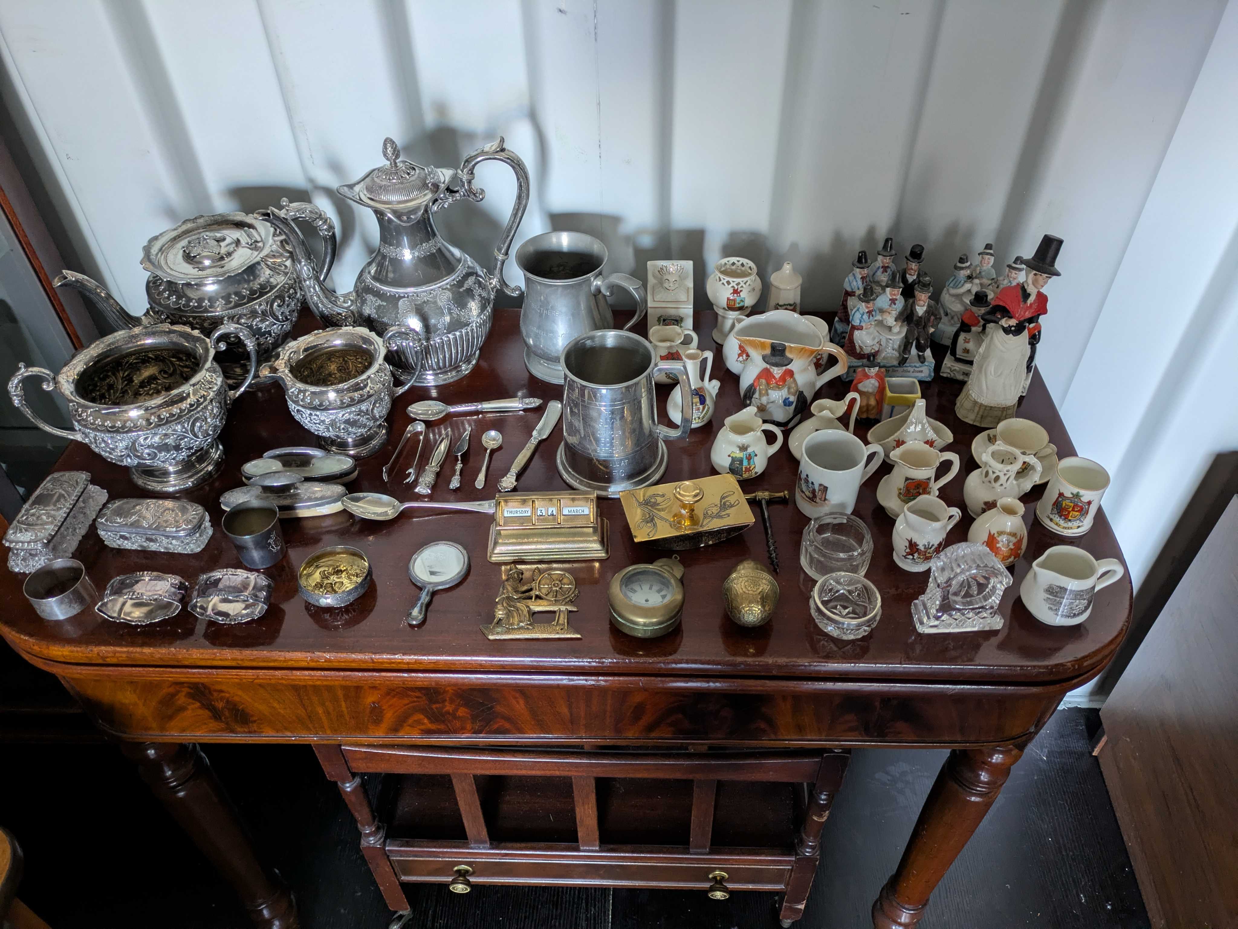 A polished table laid out with silver teapots, pewter tankards, brass ornaments, royal commemorative mugs and ceramic figurines