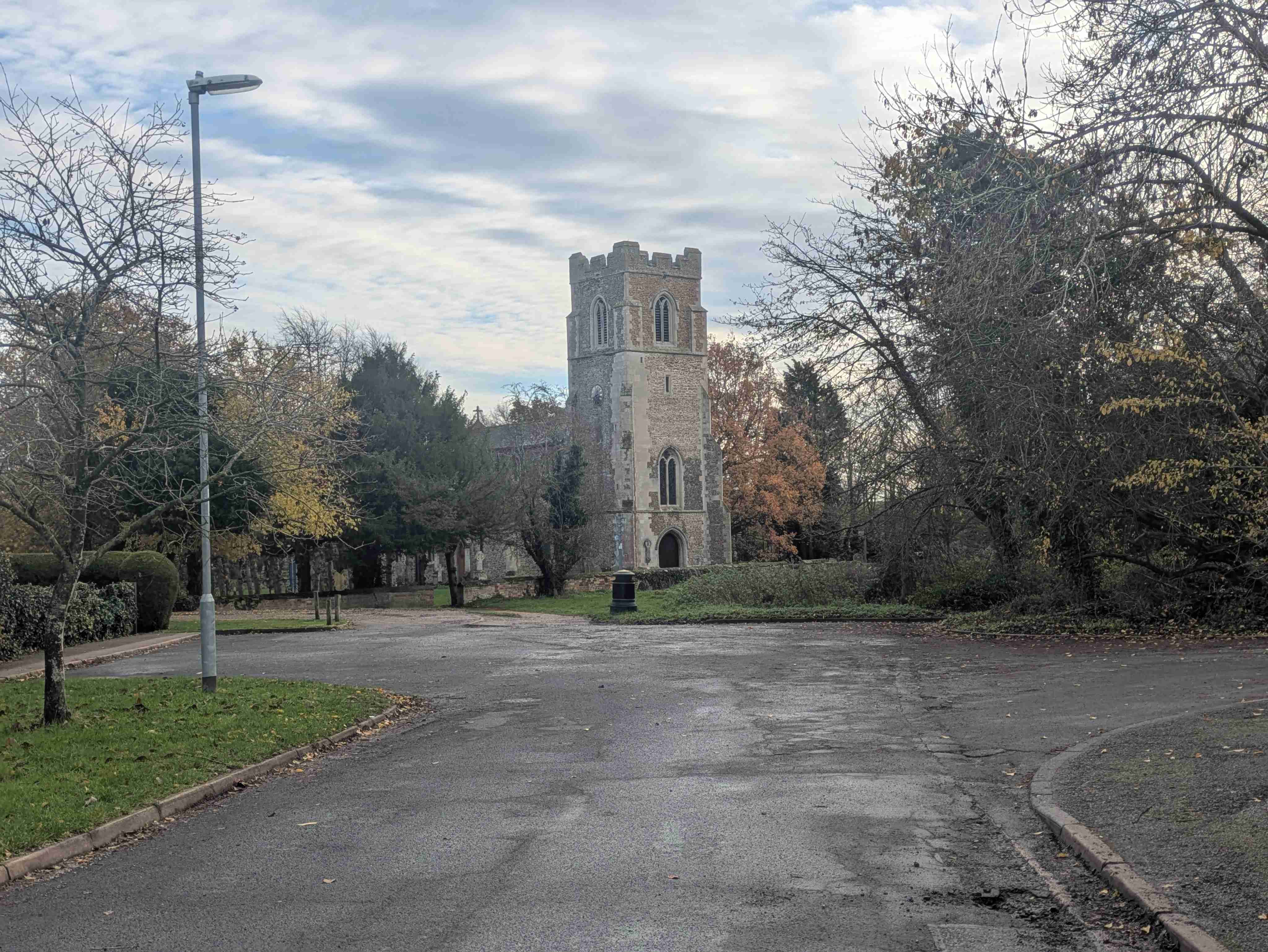 A medieval Cambridgeshire village church tower at the end of a quiet lane, autumn trees framing the stonework